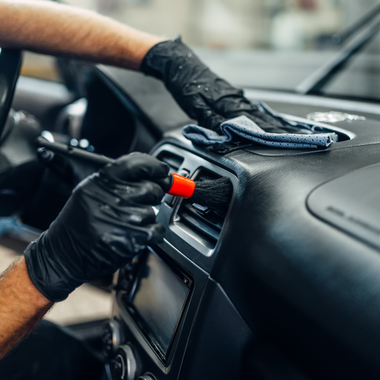 Person wearing black gloves cleaning a car dashboard with a brush and cloth.