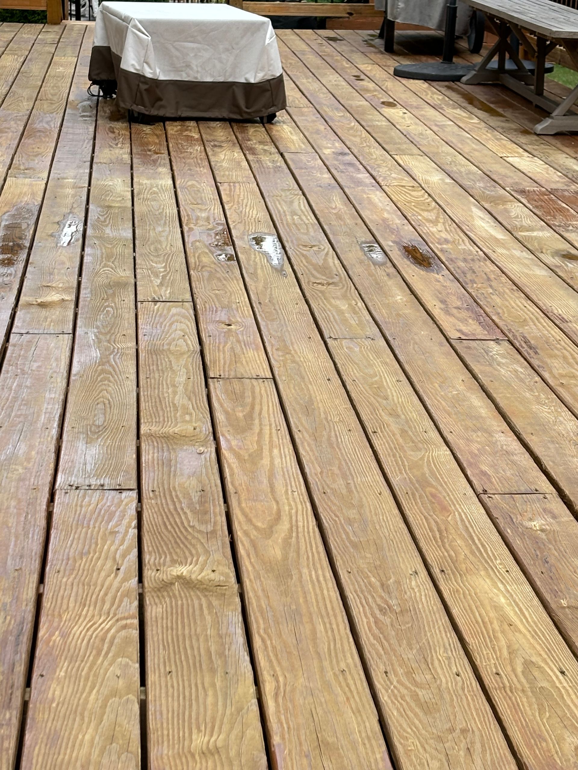 Wooden deck with wet boards, partial view of an outdoor grill covered in a light-colored cover.