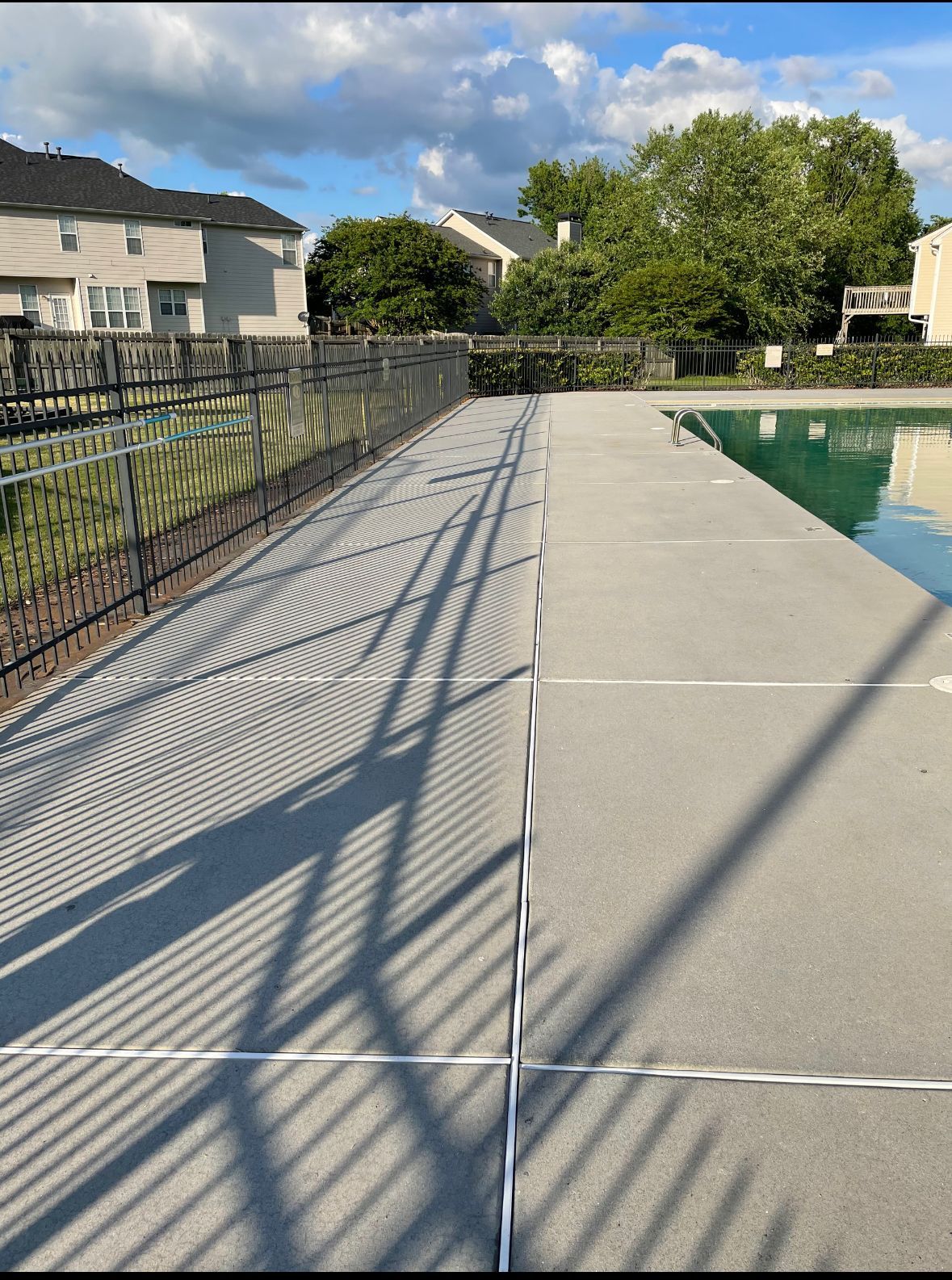 Pool deck with fence casting shadows; houses and trees in background under a partly cloudy sky.
