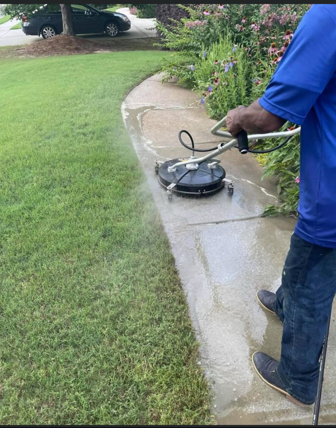 Person pressure washing a concrete sidewalk. The sidewalk is wet, and the lawn is green.