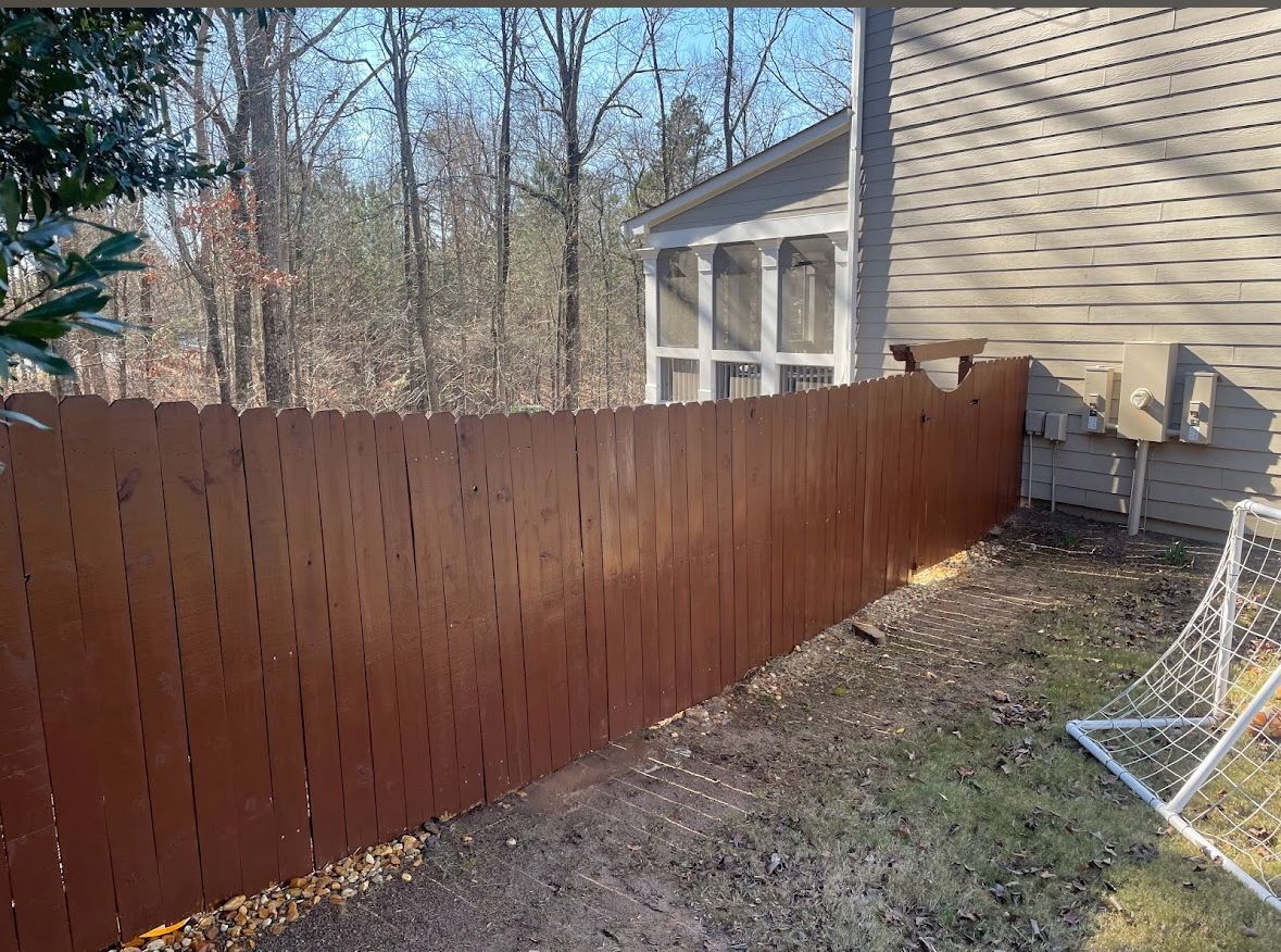 Brown wooden fence bordering a yard next to a house with bare trees in the background.