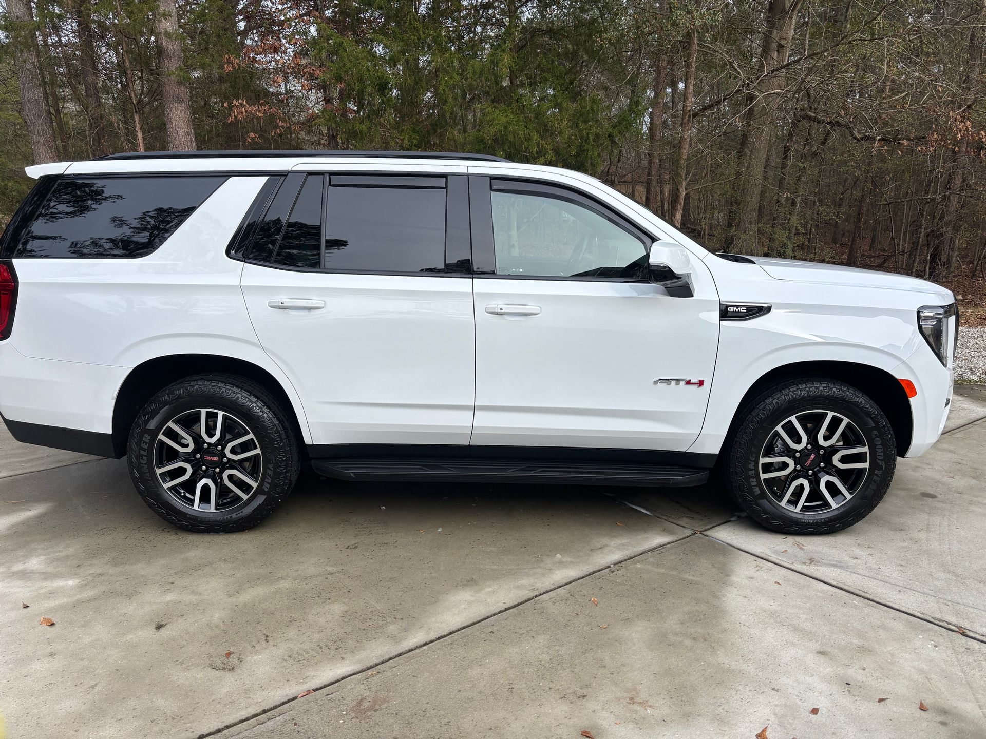 White GMC Yukon SUV parked on a concrete surface, with dark tinted windows and black and white rims, next to a wooded area.