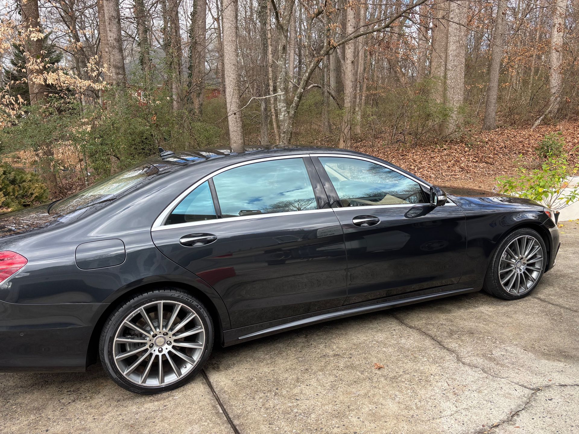 Dark gray Mercedes-Benz sedan parked on a concrete surface, tinted windows, silver rims, trees in the background.