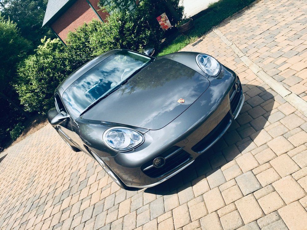 Gray Porsche Cayman parked on a brick driveway, sunny day.