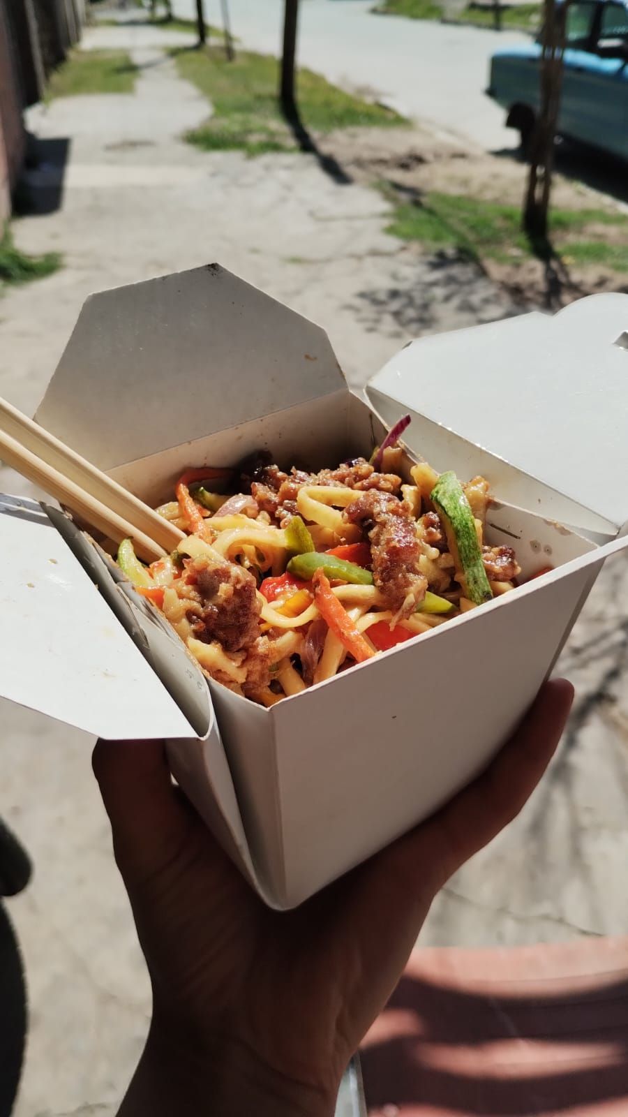 Mano sosteniendo un recipiente blanco para llevar con fideos, verduras y carne; con palillos insertados. Escenario al aire libre.