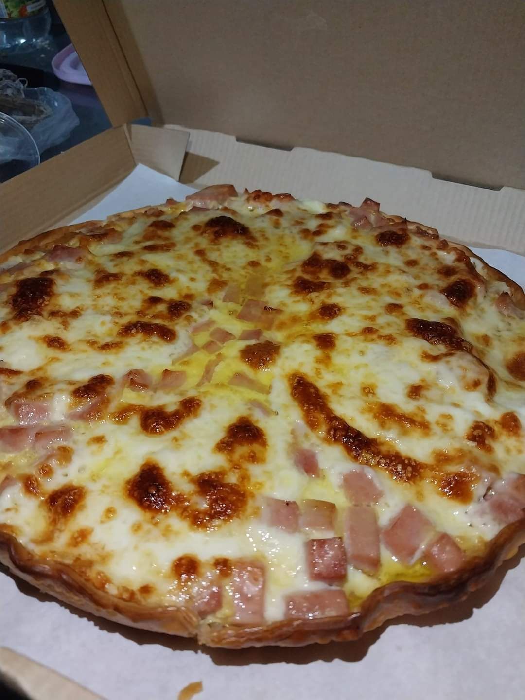 Pizza de jamón y queso en caja de cartón, con queso fundido y cubitos de jamón a la vista.