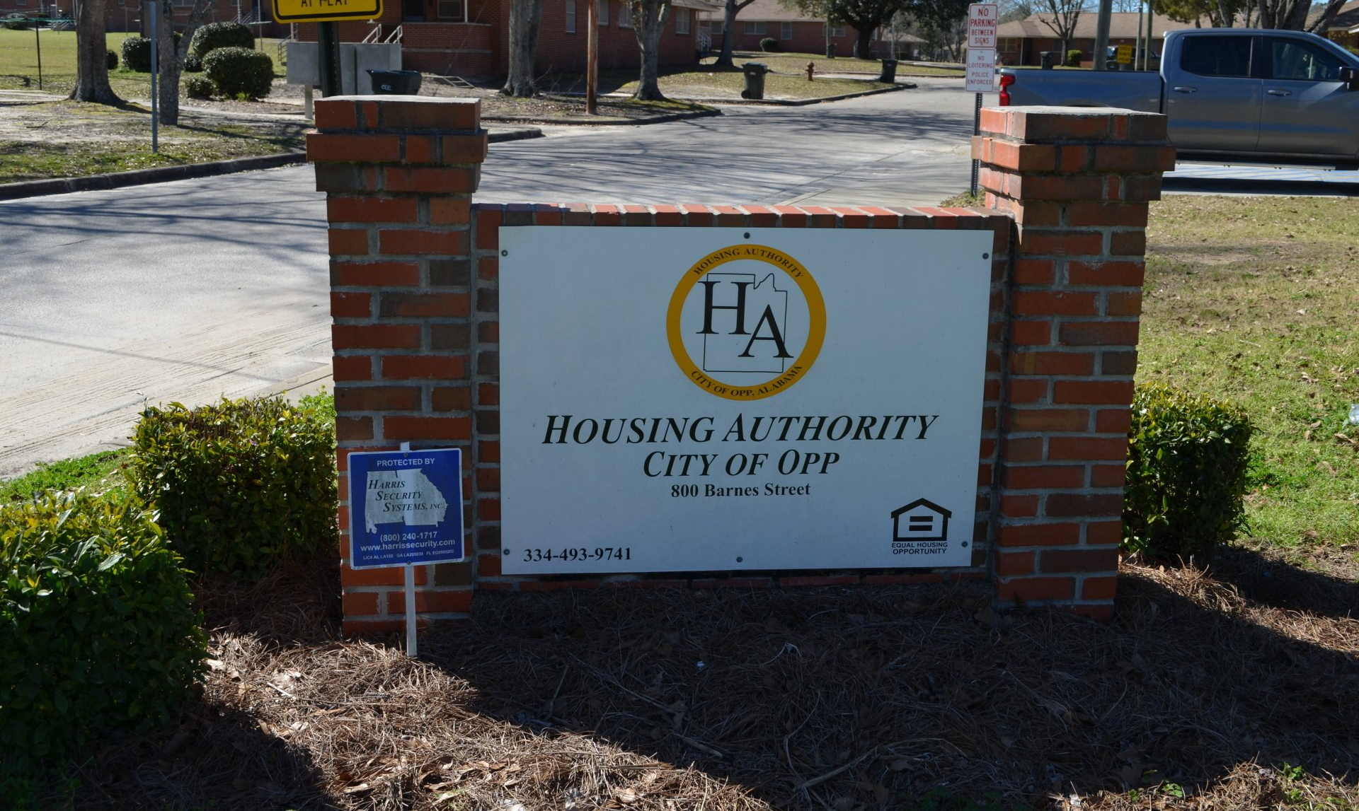 Sign for Housing Authority of Opp, City of Opp. White sign with logo between brick columns, shrubbery in front.
