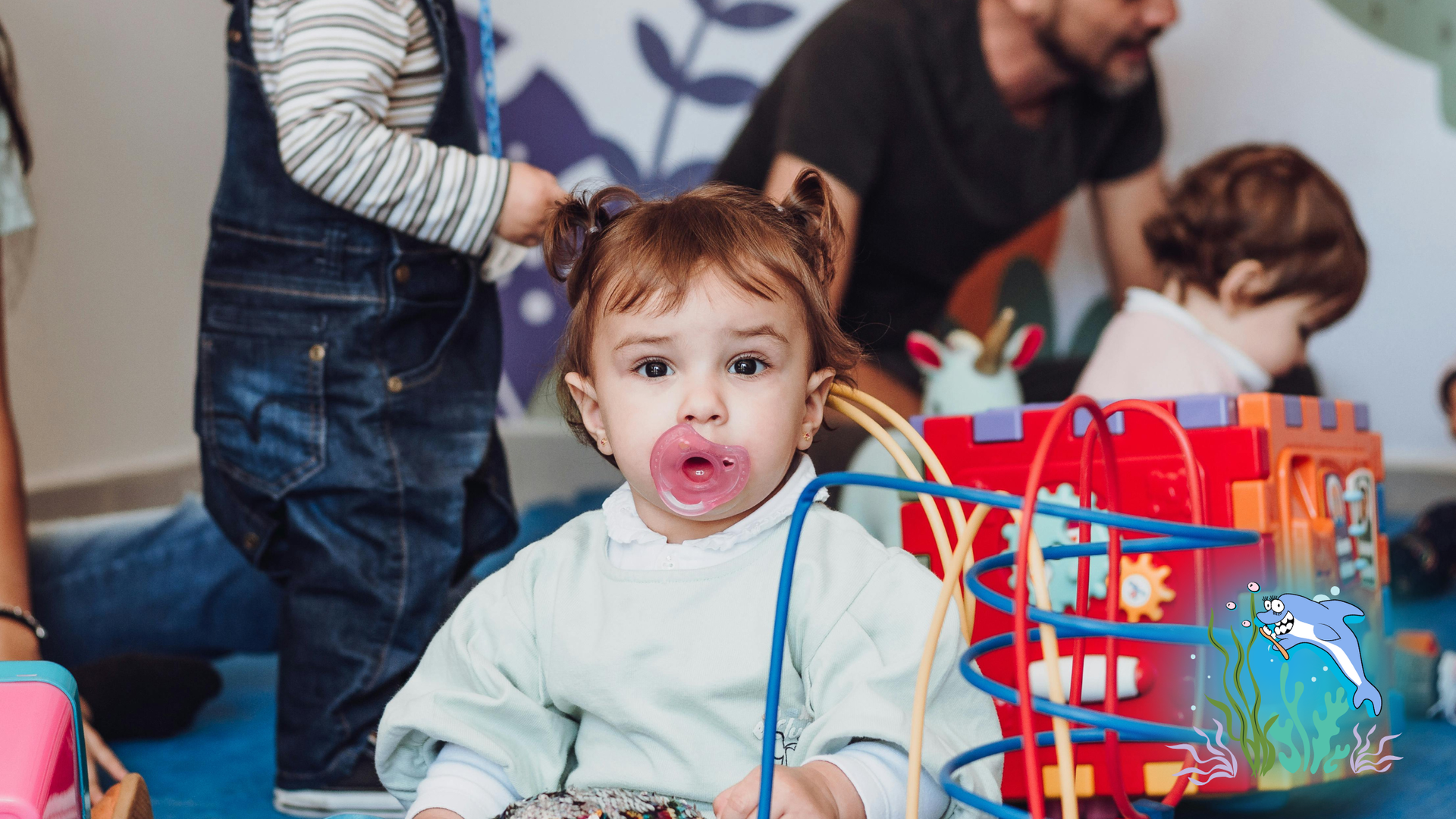 A toddler with a pacifier in a playroom stares forward, surrounded by toys and other children.