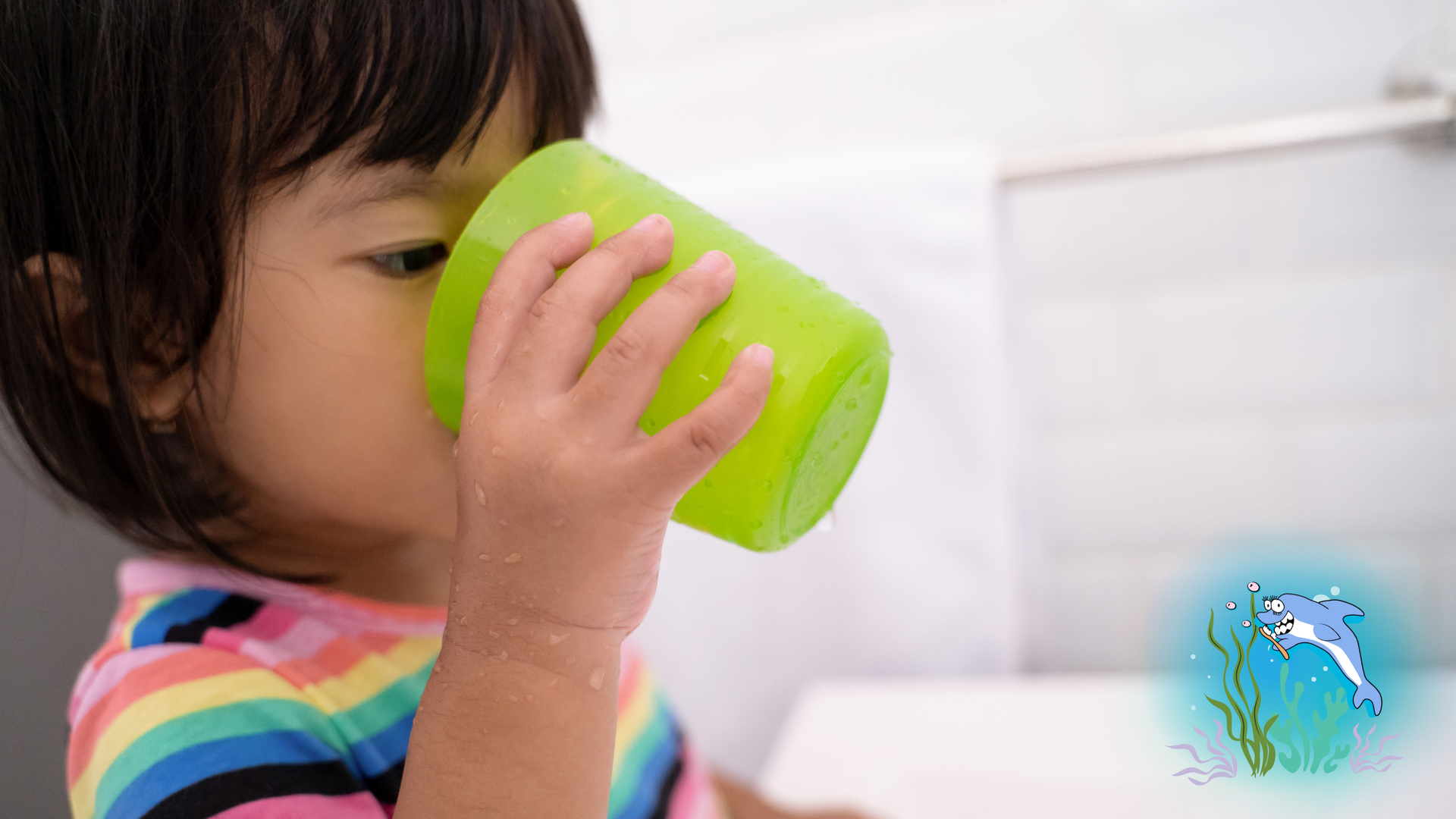 Child drinking from a green cup in a bathroom, wearing a rainbow striped shirt.
