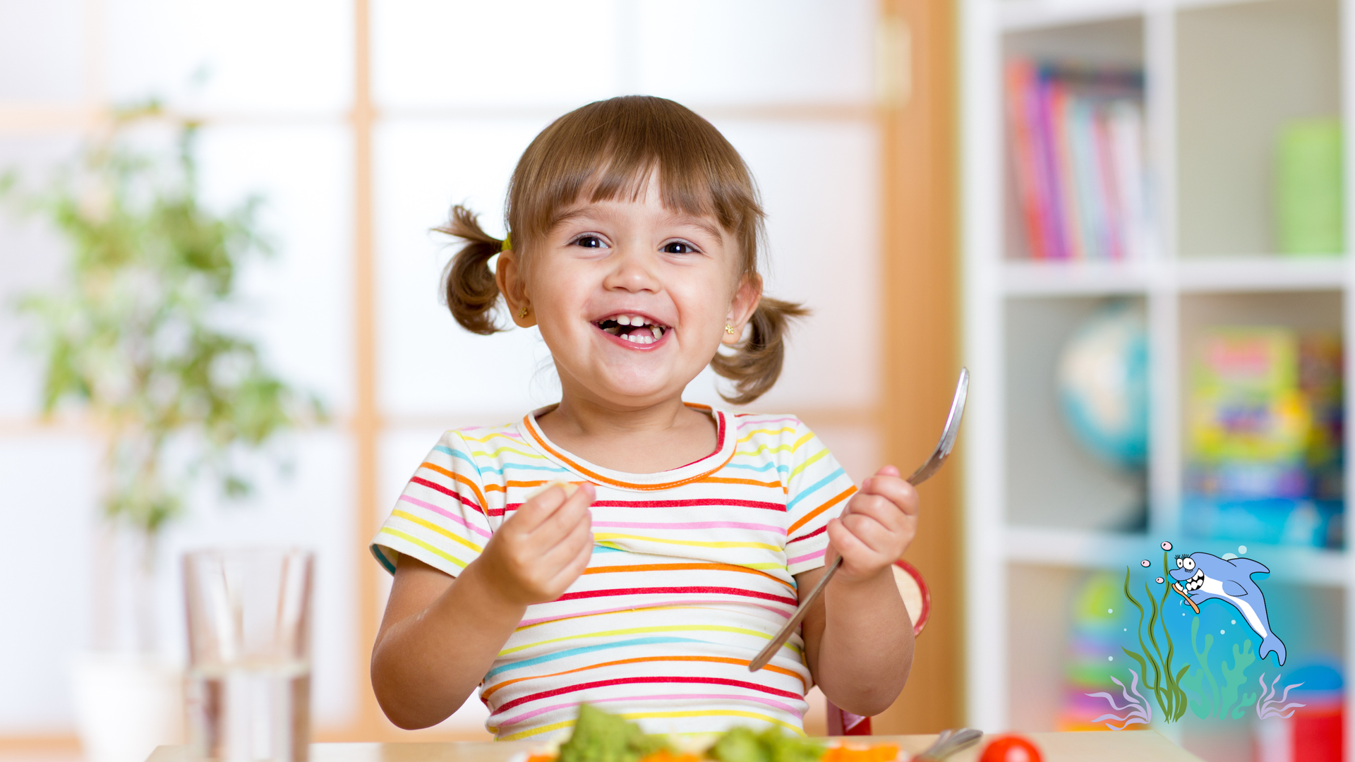Smiling child with pigtails holding a fork, eating vegetables at a table, indoors.