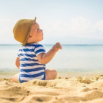 A baby is sitting on the beach wearing a hat and looking at the ocean.