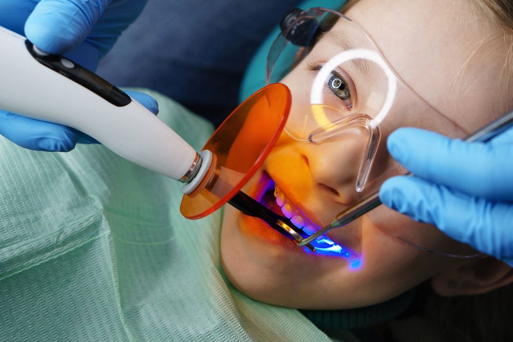 Dentist using a blue light curing tool on a patient's mouth. Patient wears safety glasses, in dental chair.