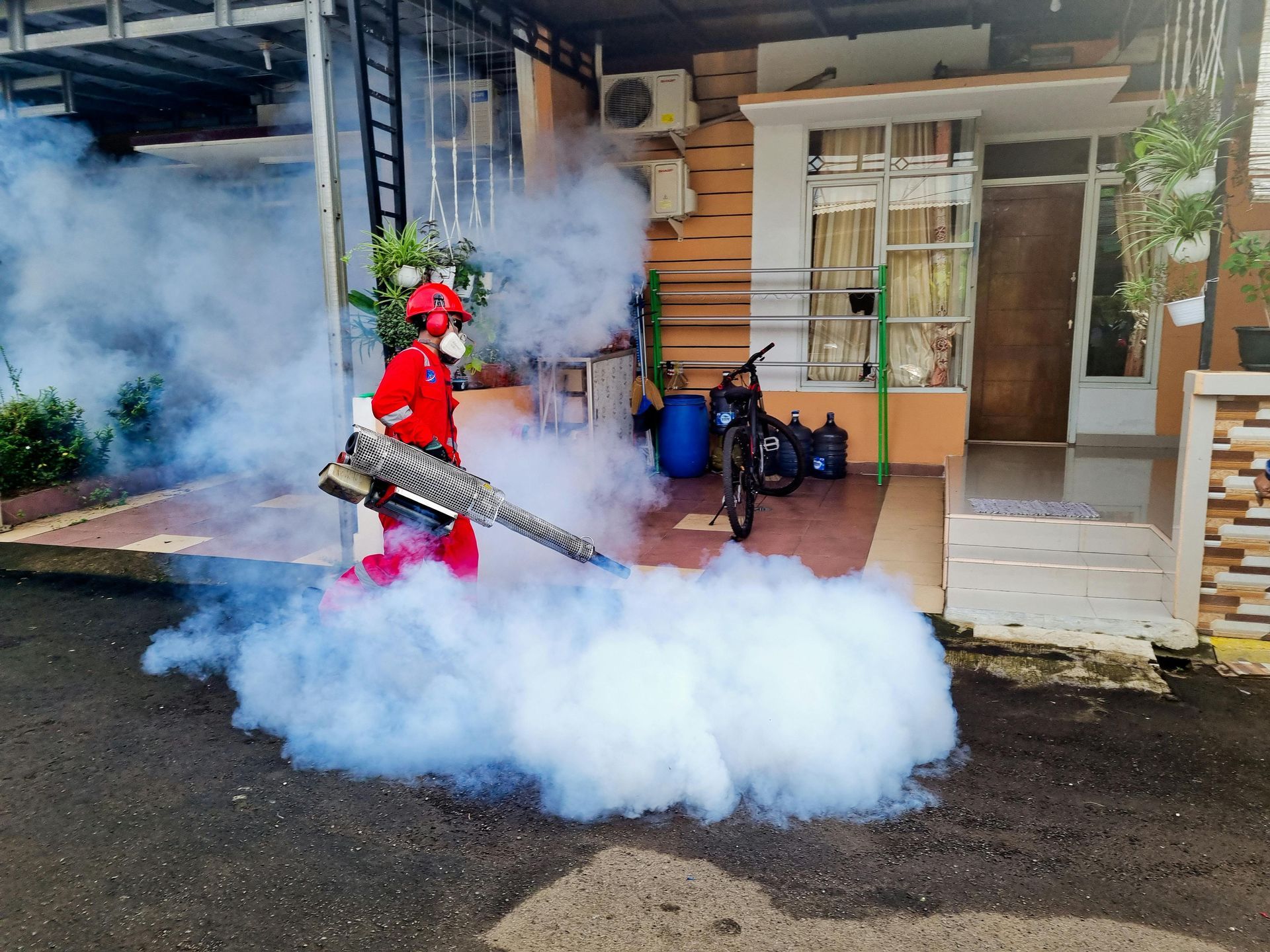 Person in red protective suit fogging a residential street with a pesticide sprayer.