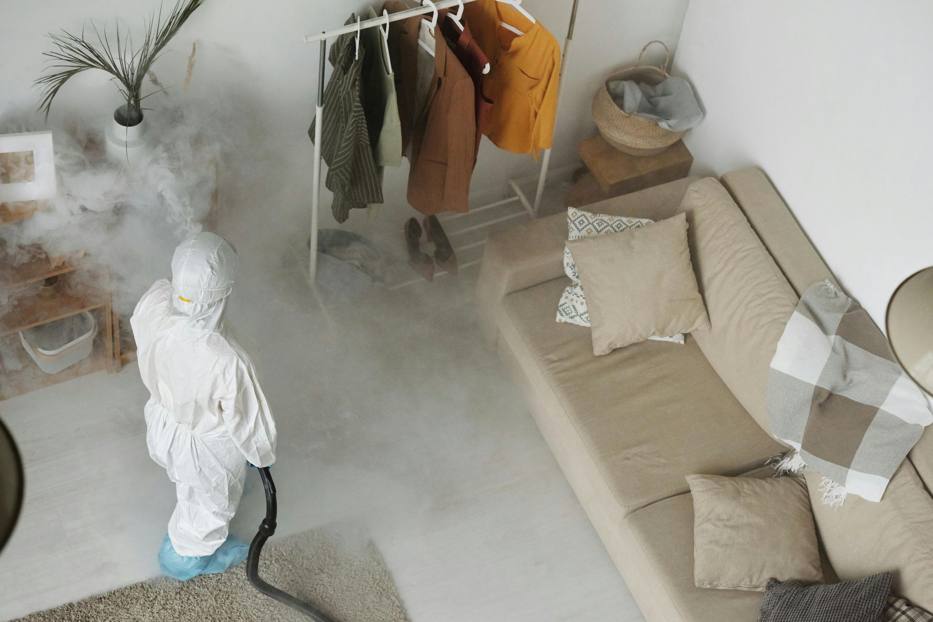 Person in hazmat suit sanitizing a room with a fogger, near a couch and clothes rack.