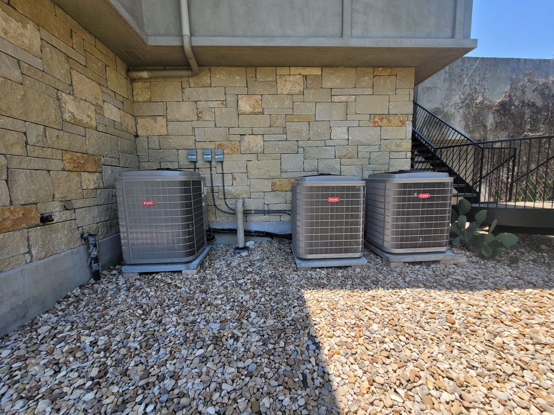 Three outdoor HVAC units beside a stone wall on a gravel patio