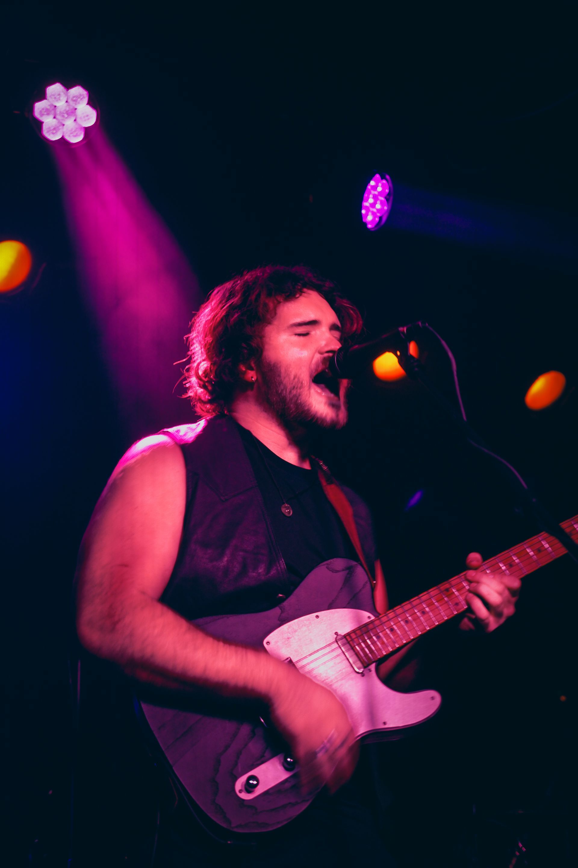 A musician with long hair sings and plays a white electric guitar on stage, bathed in purple and pink stage lights.