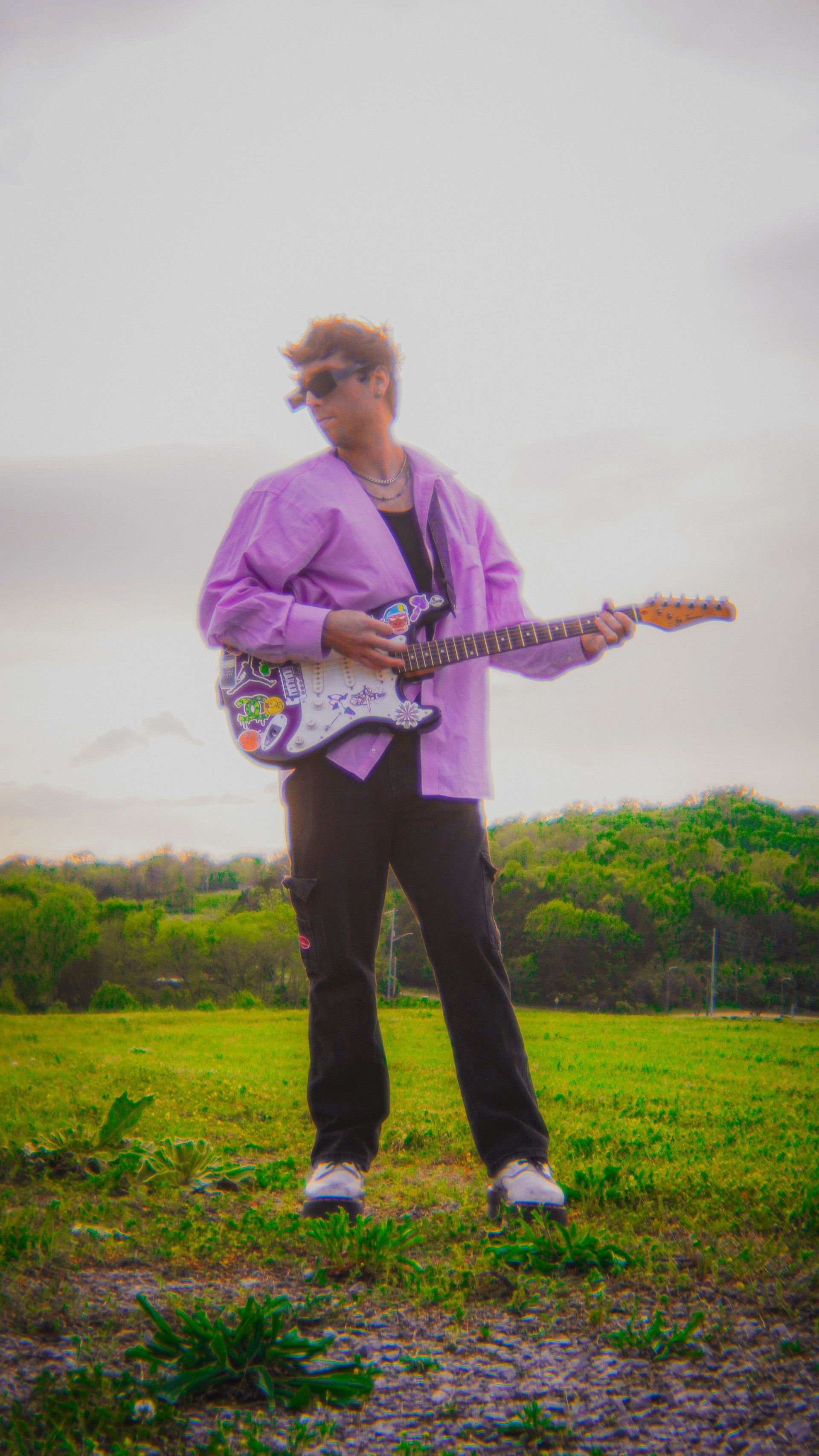 A man in a purple shirt is holding a guitar in a field.