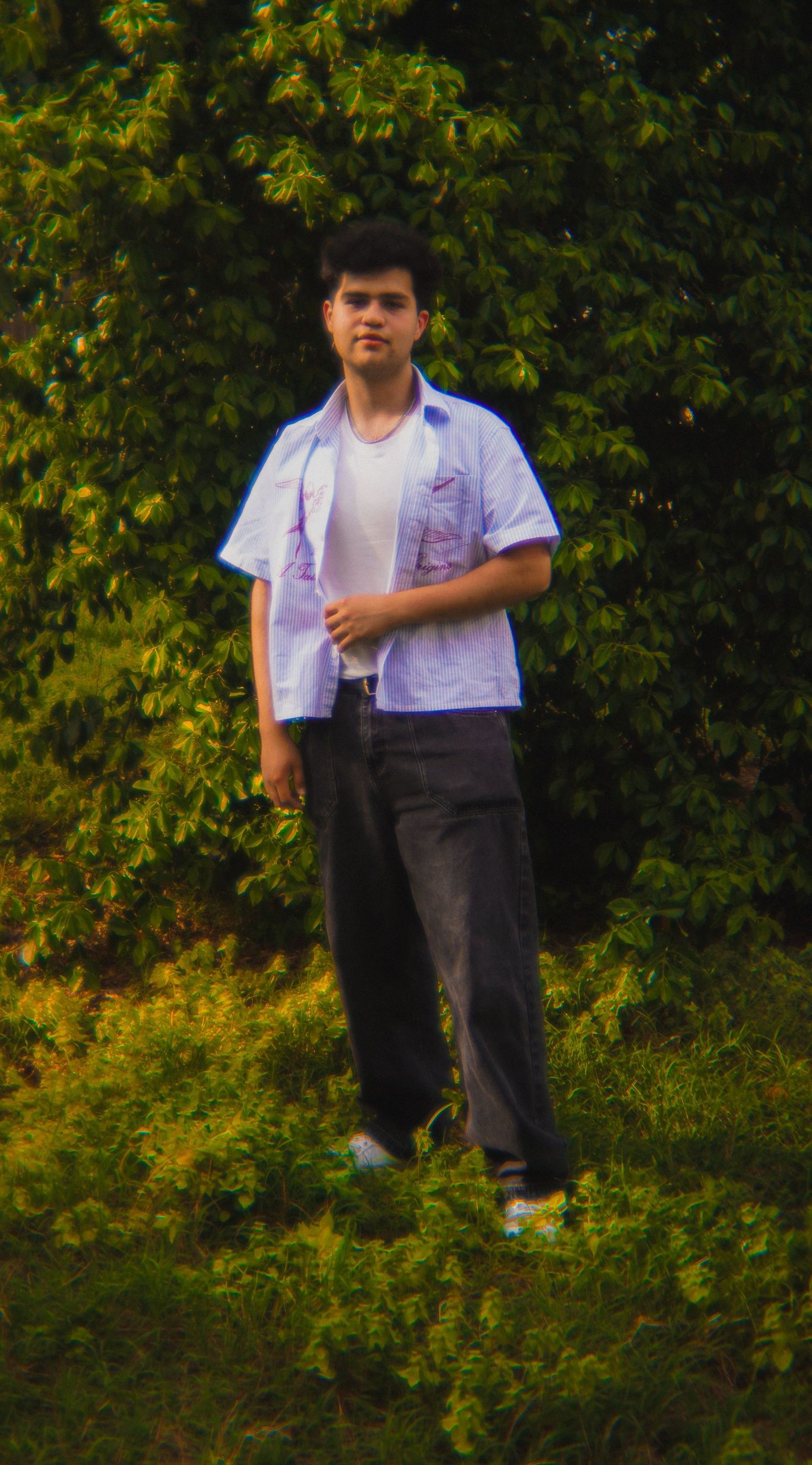 A young man is standing in the grass in front of a tree.