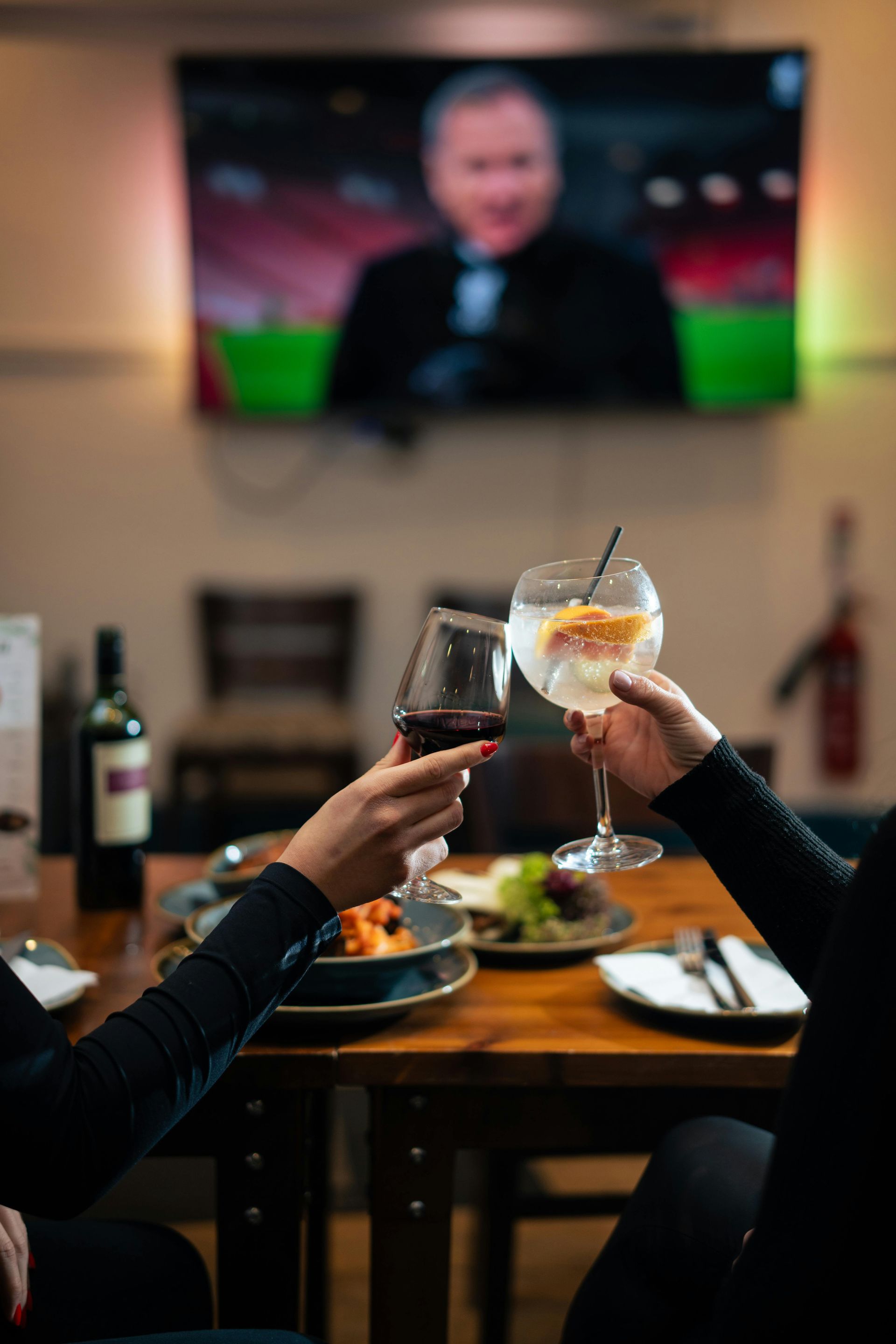 Two people are sitting at a table toasting with wine glasses in front of a television.