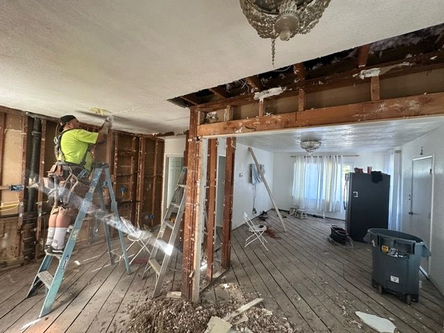 Man on ladder demolishing wall in a room. Exposed studs, wooden floor, debris scattered.