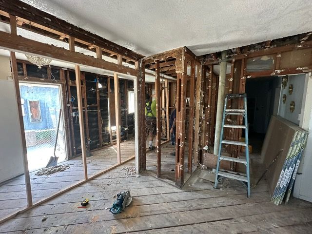 Interior room undergoing renovation, with exposed studs, debris, and ladder visible.