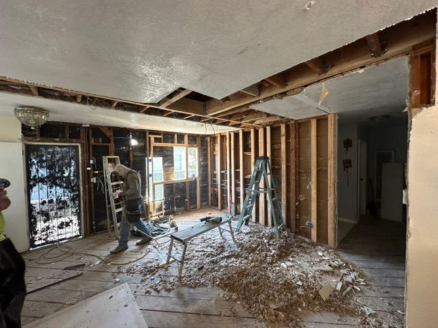 Interior demolition of a room; walls and ceiling opened. A worker is using a power tool, debris on floor.