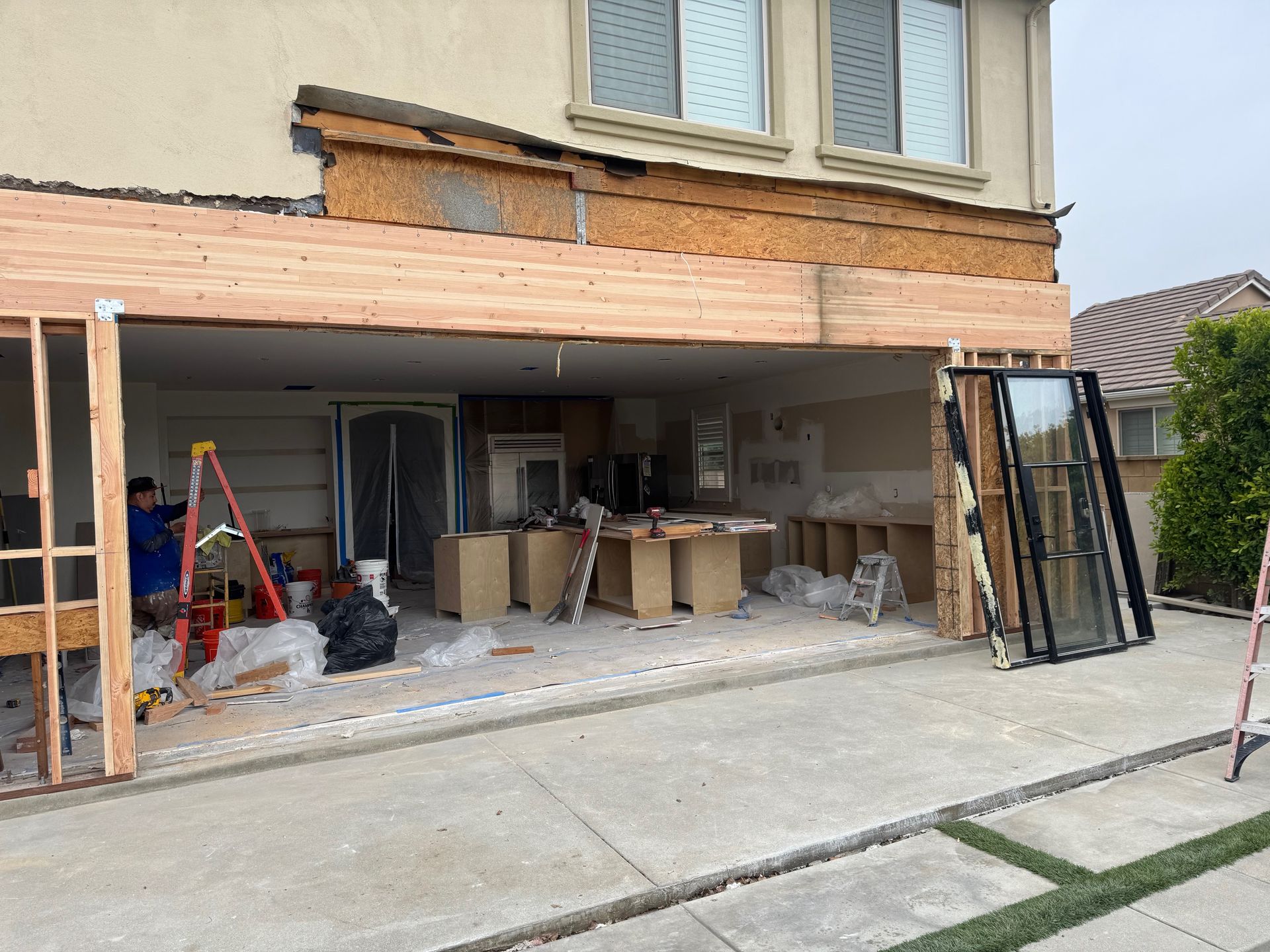 Construction of an outdoor kitchen. Exposed framing, cabinets, and a worker on a ladder visible. House exterior in background.