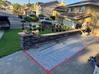 A stone wall being built in front of a house, with landscaping materials and workers nearby.
