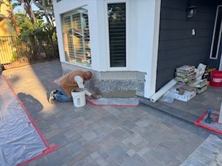 Person installing stone veneer on a building's exterior. They are kneeling, applying mortar to a wall near a window.