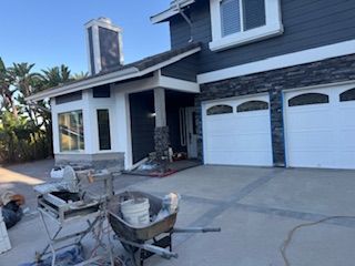 House exterior under construction, with dark siding, white trim, and stone accents. Tools and materials are visible in the driveway.