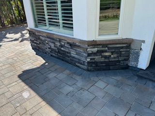 Corner of a building with stone veneer base and window with shutters, next to a paved patio.