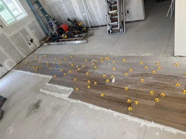 Floor tile installation in progress; wood-look tiles are laid with yellow leveling spacers, concrete floor and unfinished walls visible.