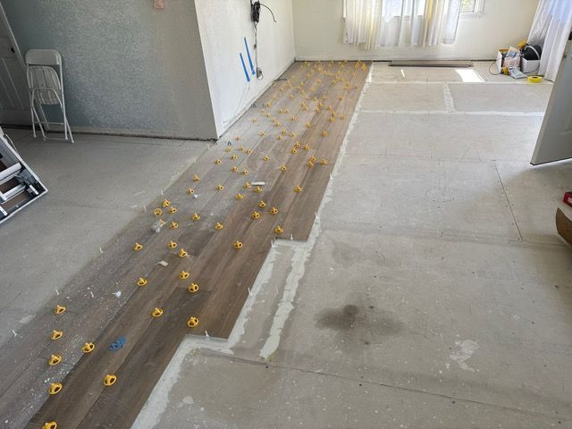 Interior room under renovation, with wood-look tile being laid on a concrete floor. Yellow spacers are visible.