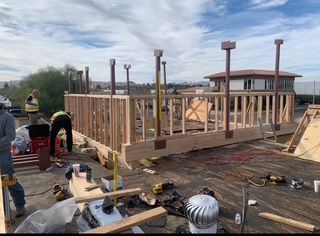 Construction workers building a wooden deck on a rooftop. Bright sky, tools, and lumber visible.