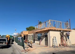 Building under construction with wooden framing, workers, and scaffolding against a clear blue sky.