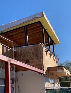 Building under construction with exposed wood framing, metal beams, and exterior sheathing against a blue sky.