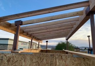 Wooden pergola under construction, with horizontal beams and brown supports against a blue sky.
