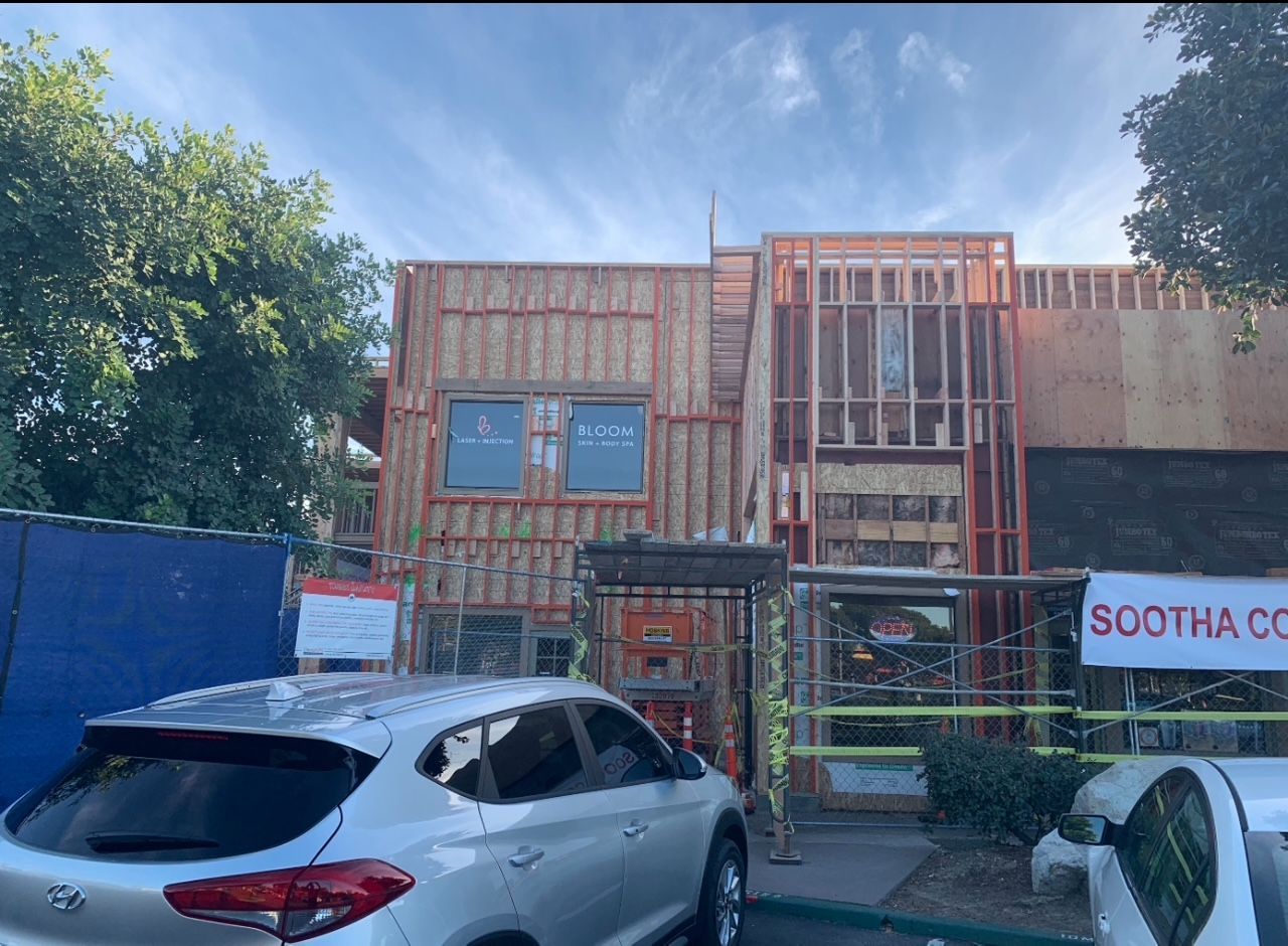 Construction site: wooden framework of a building with windows, car parked in front, trees in the background.