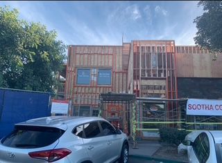 Construction site: building with exposed framing and shipping containers, cars parked in front, blue sky.