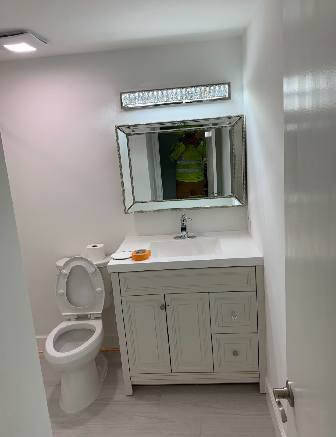 Bathroom with white vanity, mirror, toilet, and overhead light. Man in reflective vest visible in the mirror.