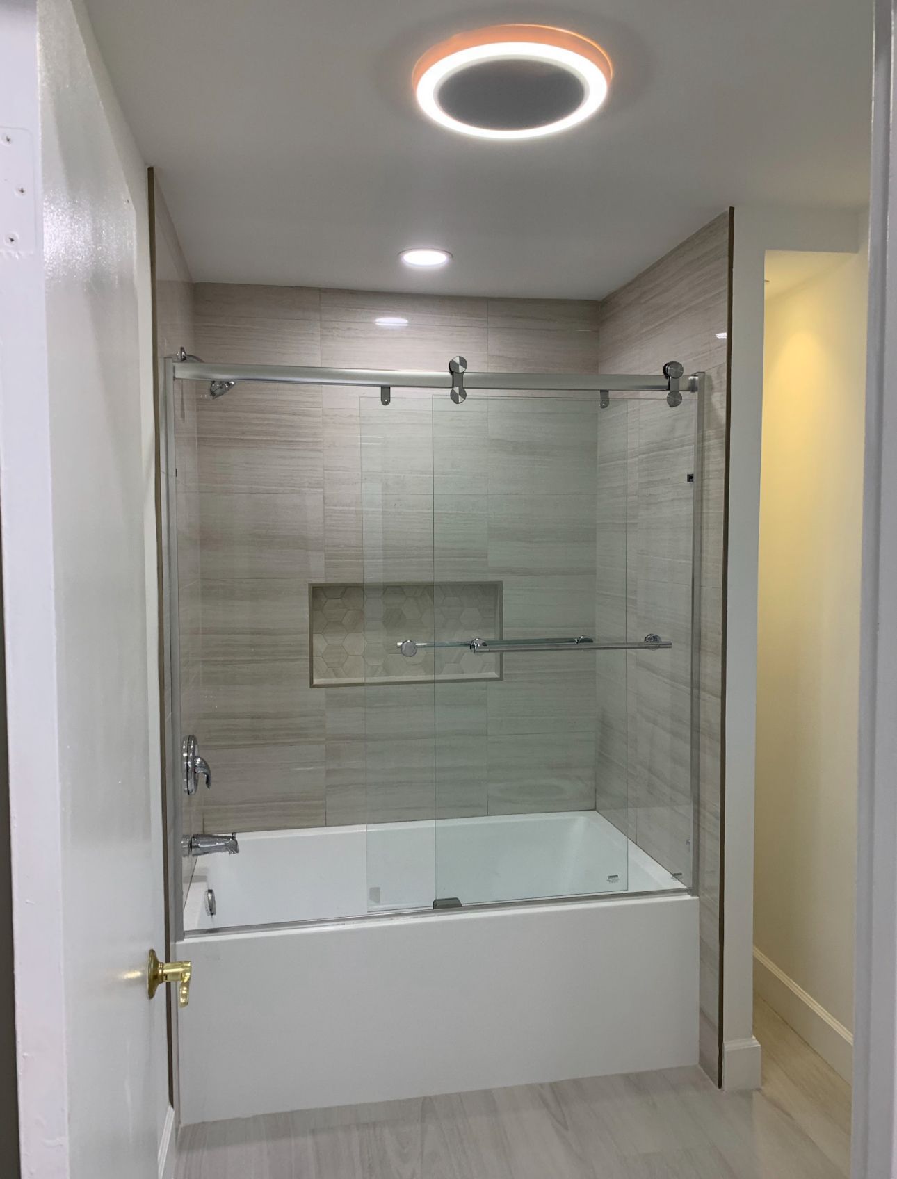 Bathroom with a white bathtub, glass shower door, and gray tiled walls. Recessed lighting.