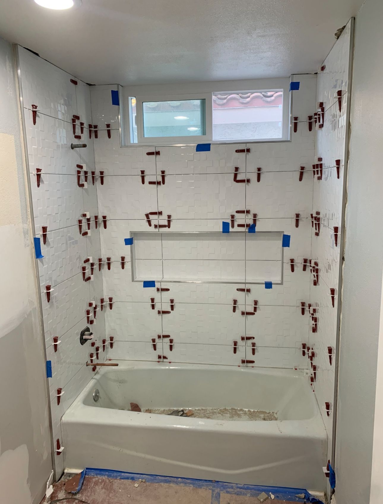 Bathroom tile installation in progress: white tiles with brown spacers, bathtub, window, and niche.