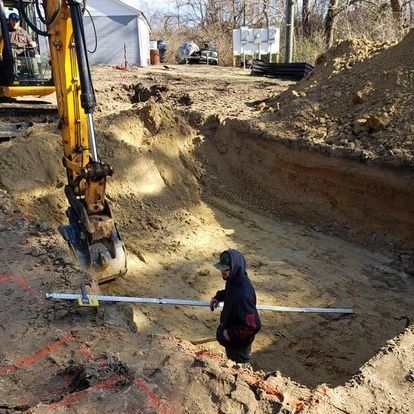 A man is measuring a hole in the ground next to a yellow excavator.