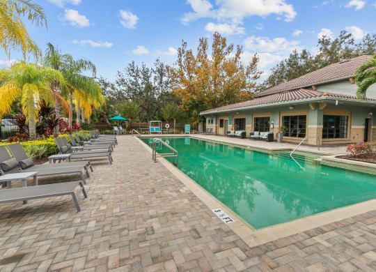 A swimming pool surrounded by lounge chairs and a building with a tile roof. Green water and palm trees are also visible.