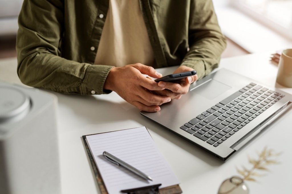 Uma pessoa sentada em umacadeira de frente para uma mesa com notebook  usa o celular com as mãos 