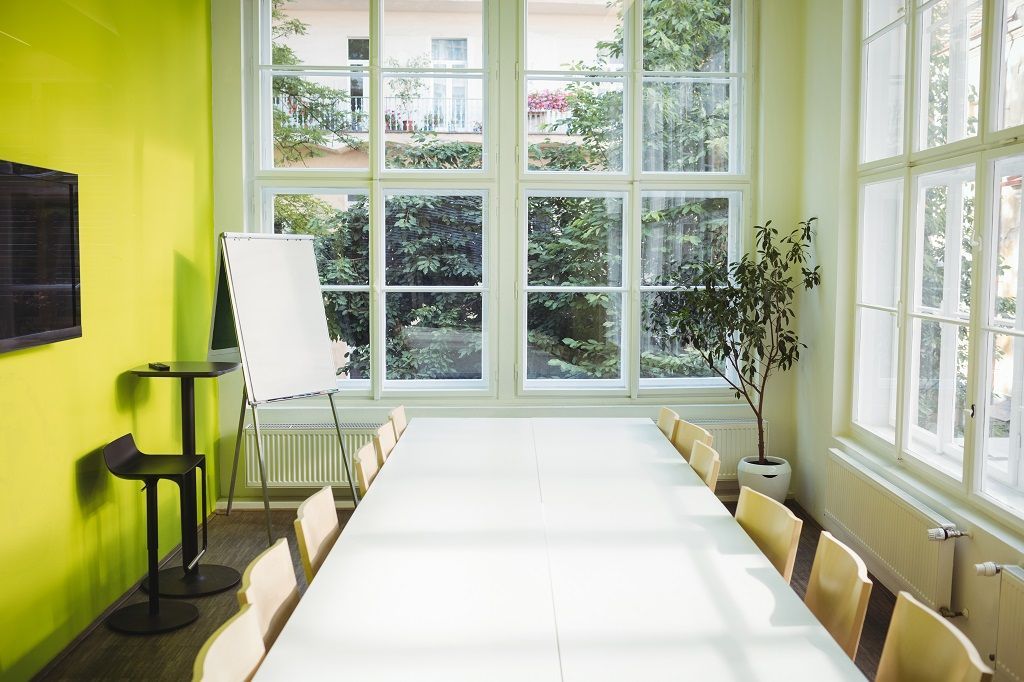 Sala de aula com uma mesa e cadeiras brancas, uma parede amarela, janelas grandes de vidro