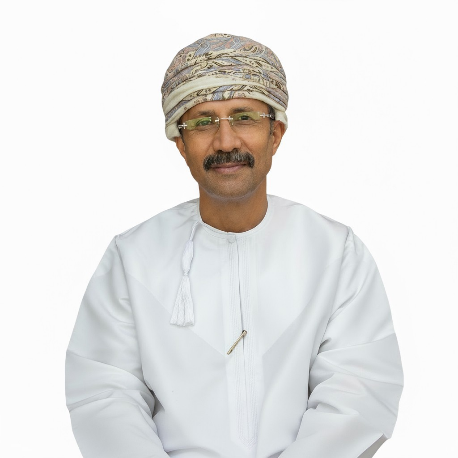 Man in white traditional Omani attire, headscarf, seated at a desk with hands clasped.