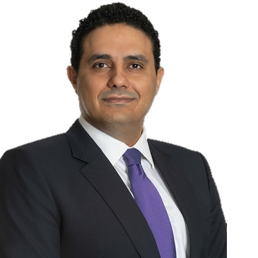 Man in white traditional Omani attire, headscarf, seated at a desk with hands clasped.