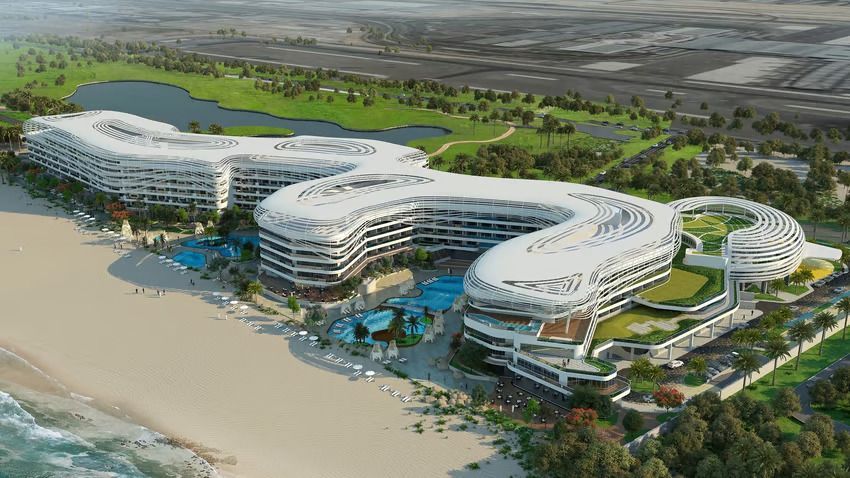 Aerial view of a modern, white, wavy hotel complex on a beach with a golf course and water features.