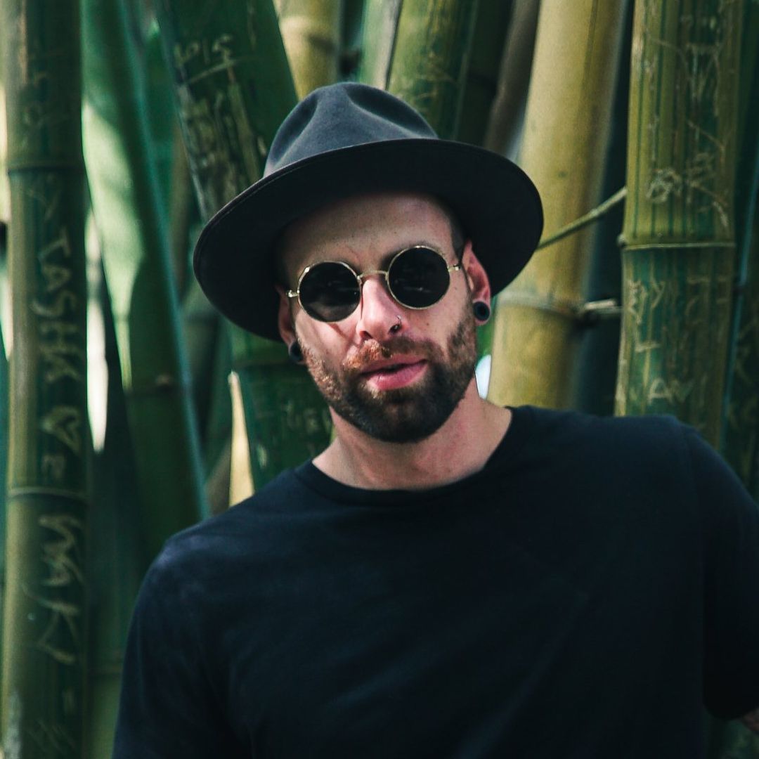 Man with beard, sunglasses, and hat posing in front of bamboo.